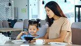 Lady shows daughter smartphone. Asian woman brunette and small kid girl watch video on black gadget sitting at restaurant table waiting for order close view