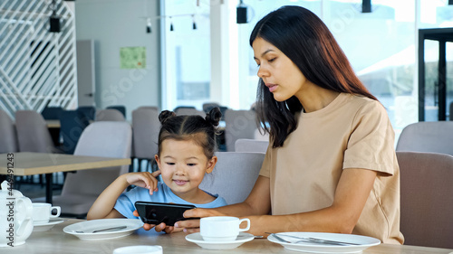 Lady shows daughter smartphone. Asian woman brunette and small kid girl watch video on black gadget sitting at restaurant table waiting for order close view