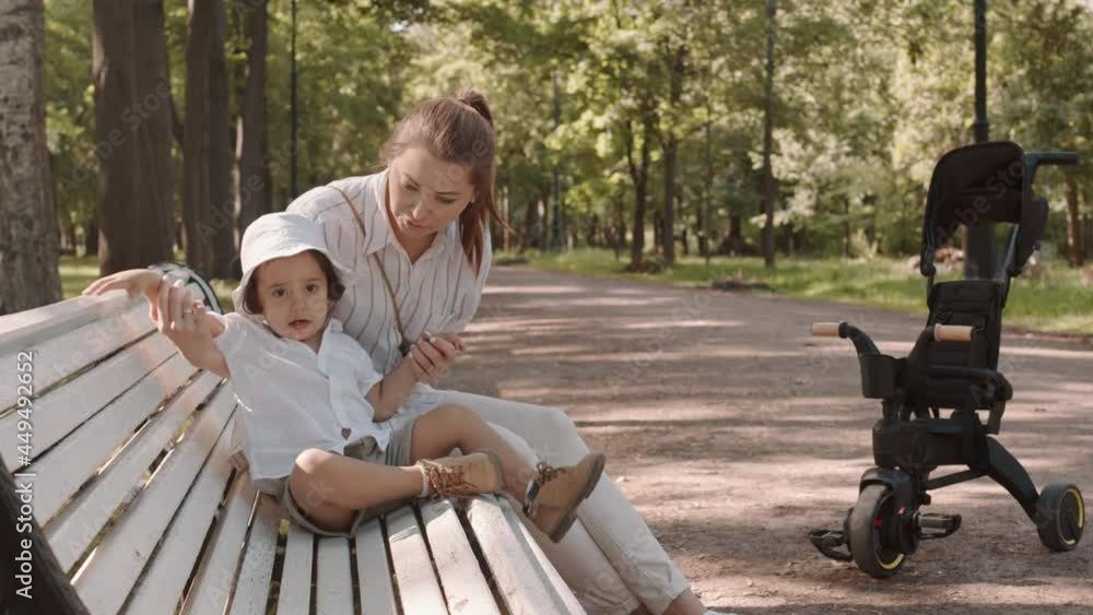 Medium shot of young woman and her cute toddler son sitting on wooden bench in park playing, enjoying summer together Medium shot of young woman and her cute toddler son sitting on wooden bench in park playing, enjoying summer together