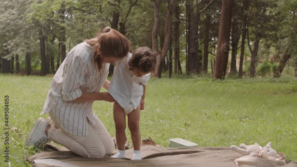 Young woman changing toddler son nappy while spending time outdoors in park on sunny day