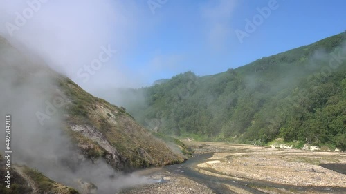 Geysers are visible in the rocky riverbed. Steam from hot springs flies over the ground. There is green vegetation and sulfur deposits on the mountain slopes. Kamchatka. Valley of geysers. A sunny day