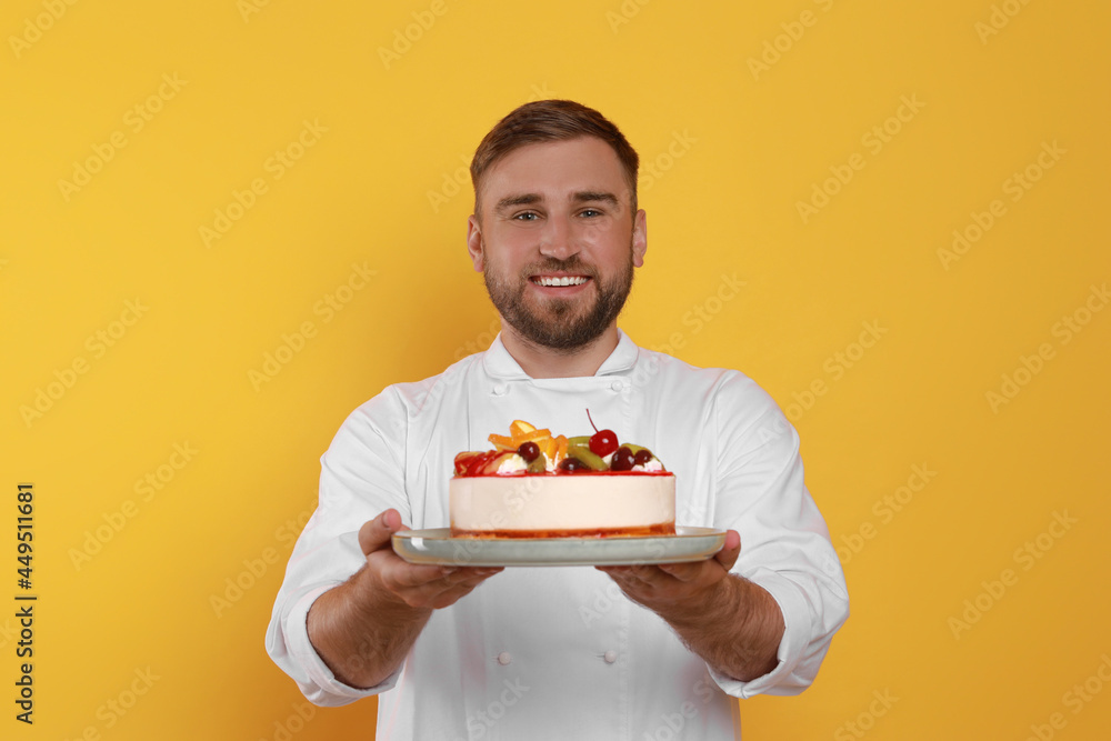 Happy professional confectioner in uniform holding delicious cake on yellow background Happy professional confectioner in uniform holding delicious cake on yellow background