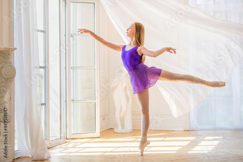 young slender ballerina in lilac leotard stands on pointe shoes in beautiful white room near a postcard window, the breeze waves the curtain.