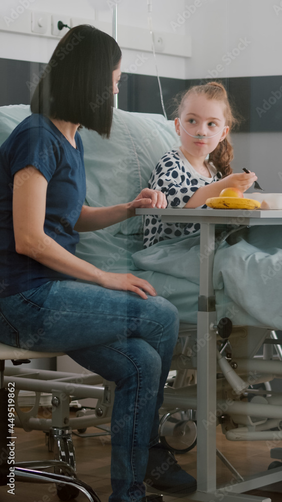 Sick child patient sitting in bed with oxygen nasal tube eating healthy food during lunch in hospital ward. Hospitalized girl recovering after medical surgery having breathing sickness