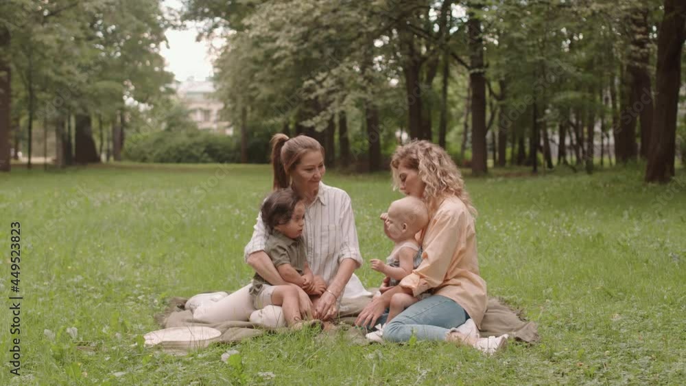 Wide shot of two happy young women and their cute little toddler children sitting on green grass in park enjoying summertime Wide shot of two happy young women and their cute little toddler children sitting on green grass in park enjoying summertime