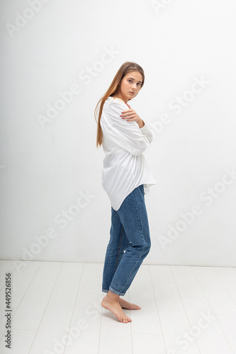 young attractive caucasian woman with long brown hair in shirt, blue jeans on white studio background. skinny pretty lady posing with bare feet at bright room. portrait of beautiful female