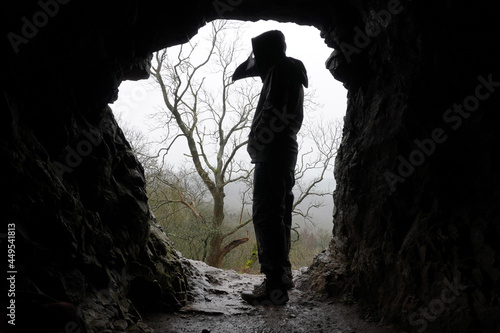 A scary, hooded figure wearing a plague doctor mask, standing in the entrance of a cave on a misty winters day