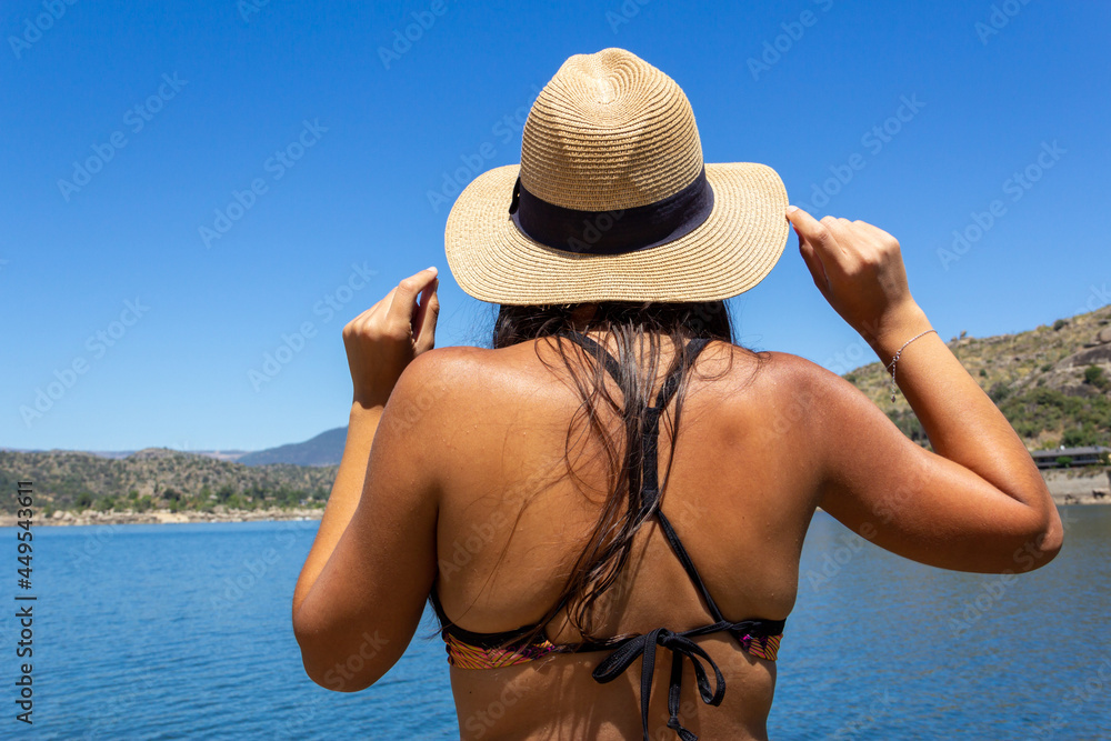 Young woman from the back in a bikini with a hat looking at a lake. Summer. Selective focus.