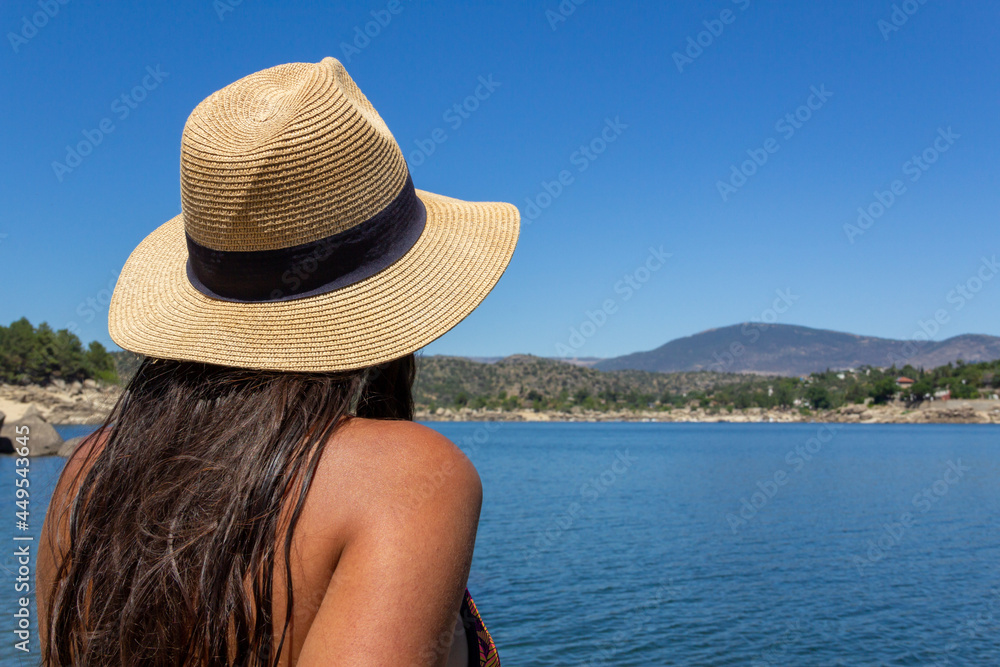 Young woman sitting on a rock, her back, in a bikini with a hat looking at a lake. Summer. Selective focus.