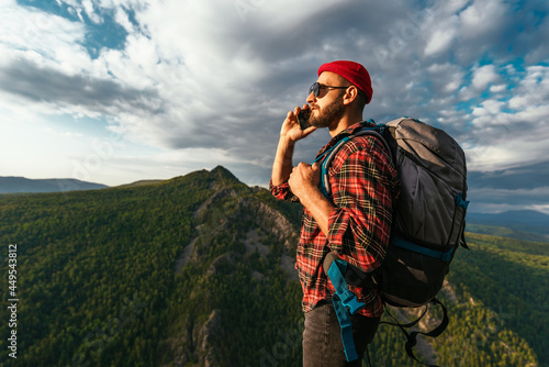 A male traveler in a red hat with a backpack is talking on the phone against the background of mountains. A man in hiking clothes talks on a smartphone at sunset in the mountains. Phone conversation