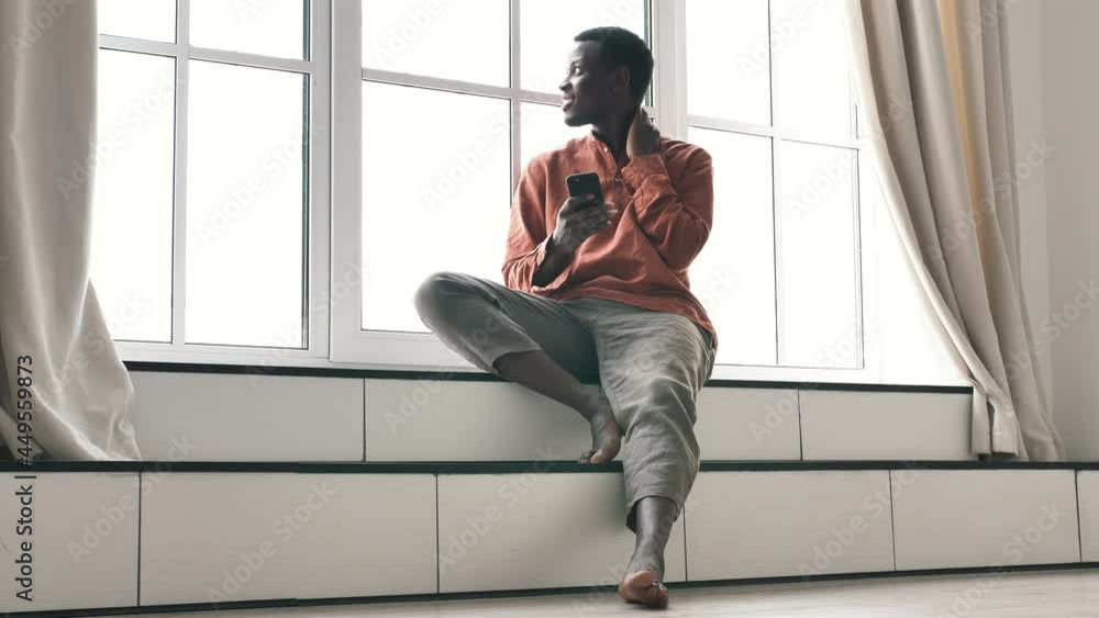 Emotional surprised African-American man in stylish casual clothes looks at smartphone screen sitting on windowsill closeup Emotional surprised African-American man in stylish casual clothes looks at smartphone screen sitting on windowsill closeup