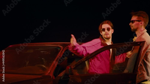 Two men in sunglasses dance vigorously outside a car on the street at night.