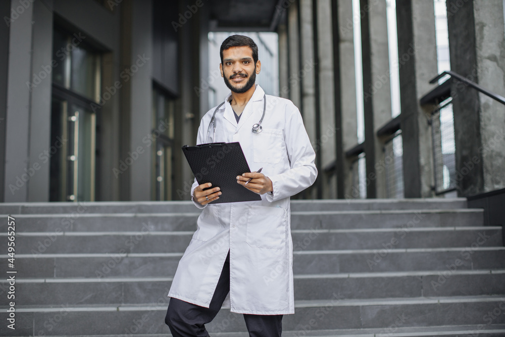 Close up front view of pleasant young Middle Eastern ethnic doctor in white gown, holding a folder, standing on stairs of modern clinic outdoors, smiling and looking at camera Close up front view of pleasant young Middle Eastern ethnic doctor in white gown, holding a folder, standing on stairs of modern clinic outdoors, smiling and looking at camera
