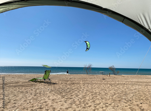 View from a tourist tent on the seashore to the shore, the sea and a male person enjoy riding kite surf board against the blue sky on a sunny day.