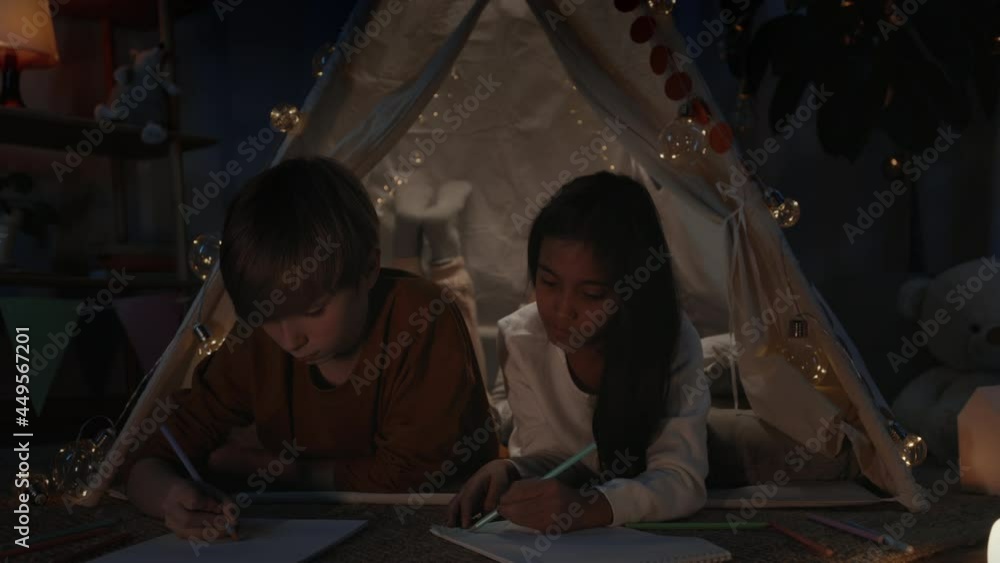 Young diverse boy and girl drawing with colored pencils while spending free time in evening. Teenagers lying on floor and talking in decorative tent while creating pictures. Young diverse boy and girl drawing with colored pencils while spending free time in evening. Teenagers lying on floor and talking in decorative tent while creating pictures.