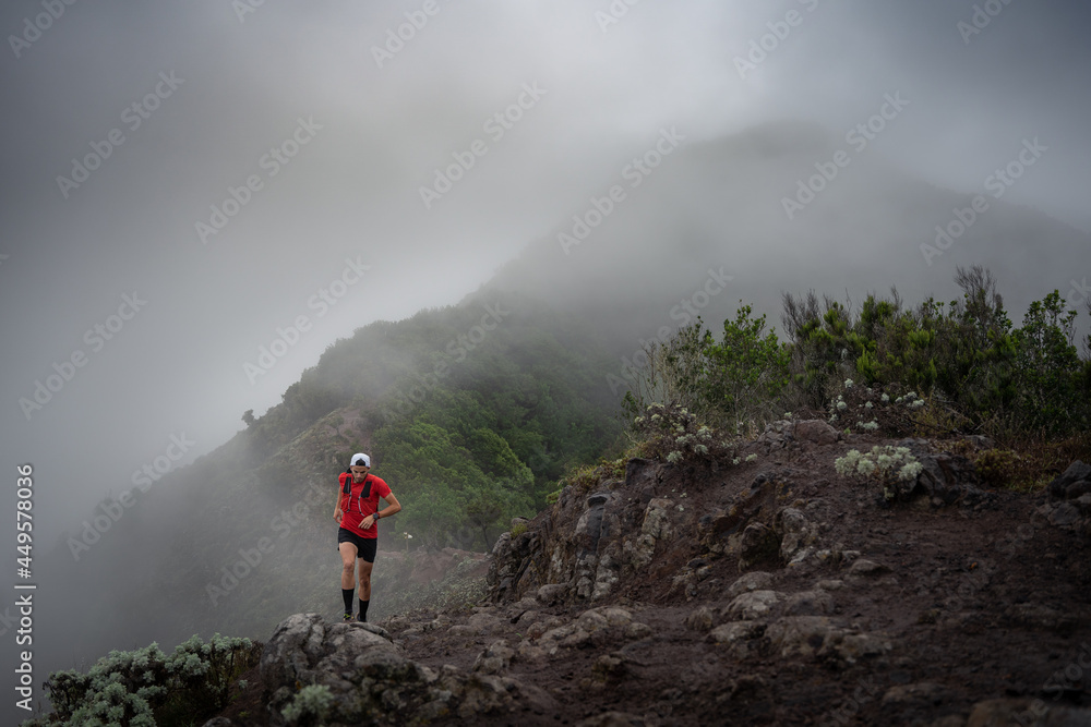 A man in a cap and red shirt doing trail running through the mountain and forest with fog and cloudy weather.