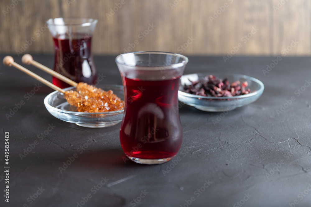Hot Karkade tea in two glasses, next to it is a glass jar with candied sugar on two wooden sticks. Hot Karkade tea in two glasses, next to it is a glass jar with candied sugar on two wooden sticks.