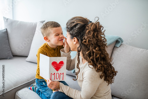 Happy little boy congratulating smiling mother and giving card with red heart during holiday celebration at home. Cheerful mother hugging son and reading handmade greeting card