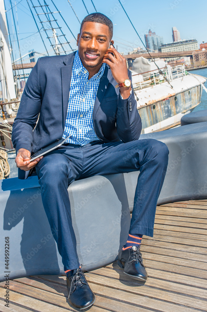 Holding a tablet computer on his lap, a young black businessman is sitting on a deck and smilingly talking on the phone. The background is a harbor.. Holding a tablet computer on his lap, a young black businessman is sitting on a deck and smilingly talking on the phone. The background is a harbor..