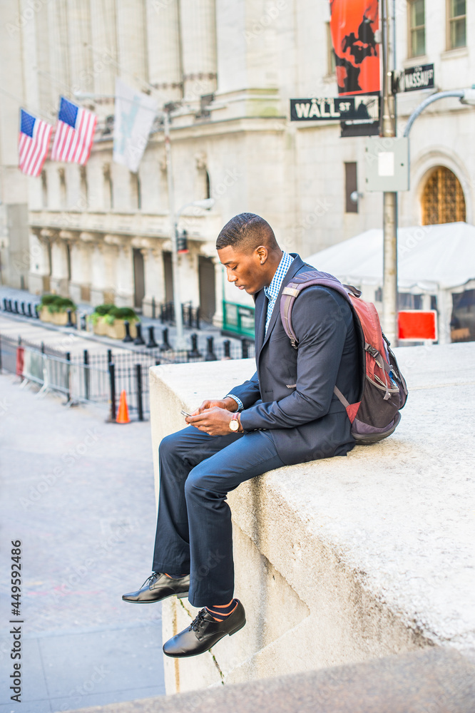Dressing in a blue suit and leather shoes, caring a backpack, a young black businessman is sitting outside to check messages on his mobile phone. There is a Wall Street sign in the background..