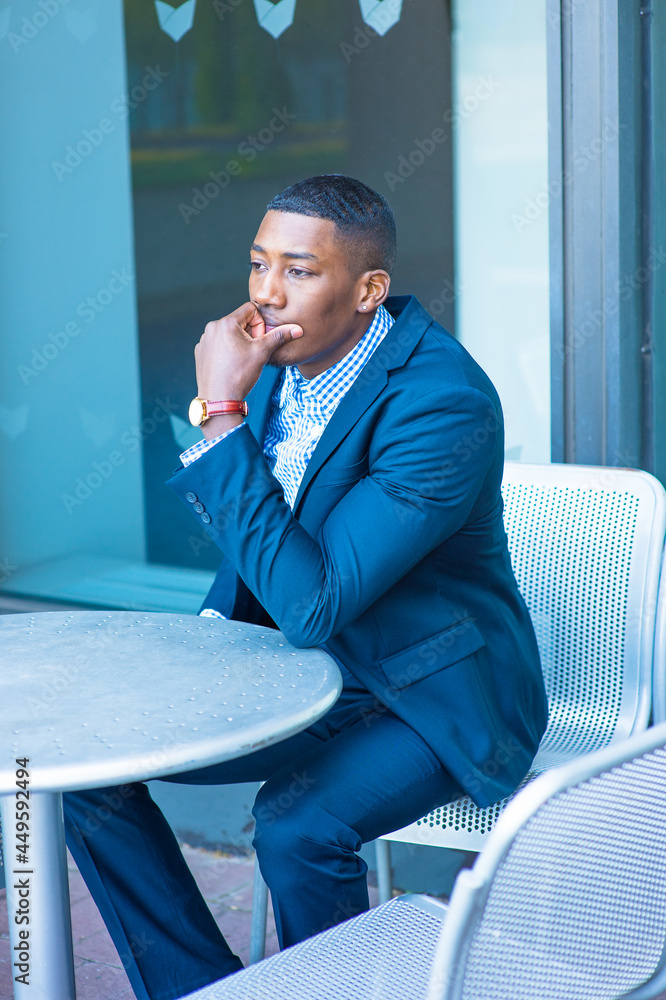 A hand touching his mouth, a young black businessman is sitting on a chair by a table outside, thinking.. A hand touching his mouth, a young black businessman is sitting on a chair by a table outside, thinking..