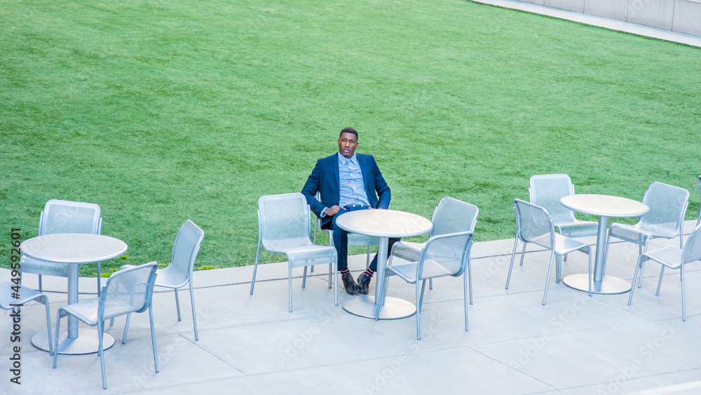 A young handsome black businessman is sitting on a chair by a green lawn and waiting for you..