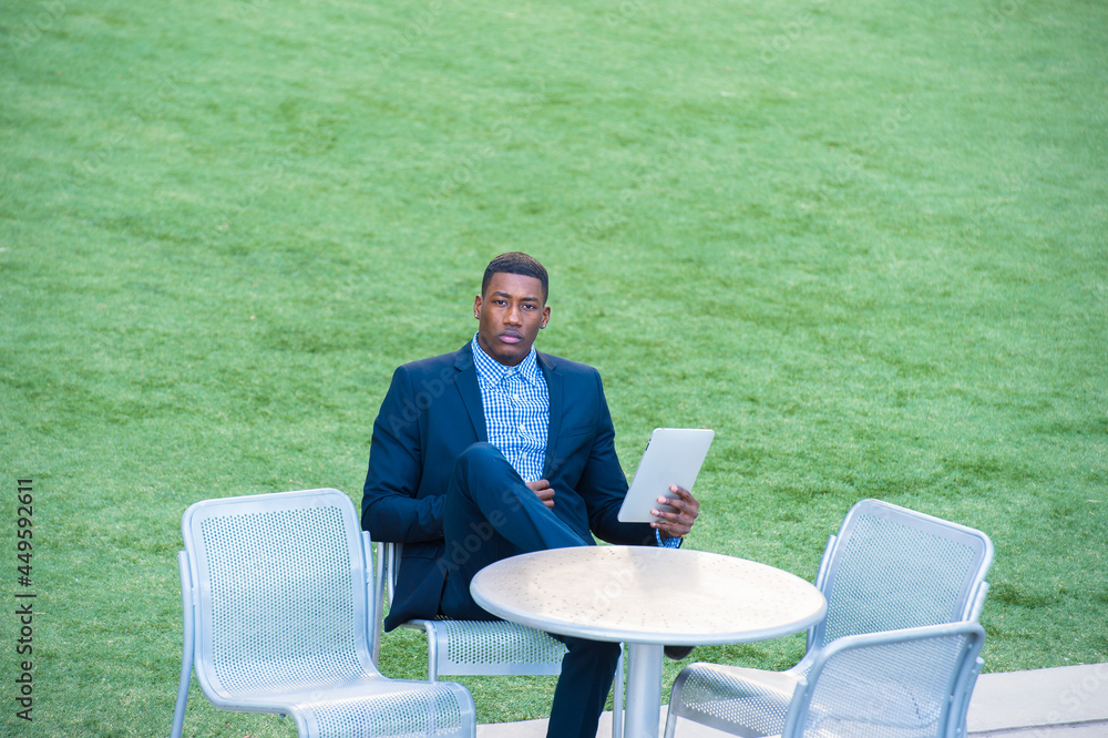 Fototapeta premium A young handsome black man is sitting on a chair by a green lawn and reading at a small computer..