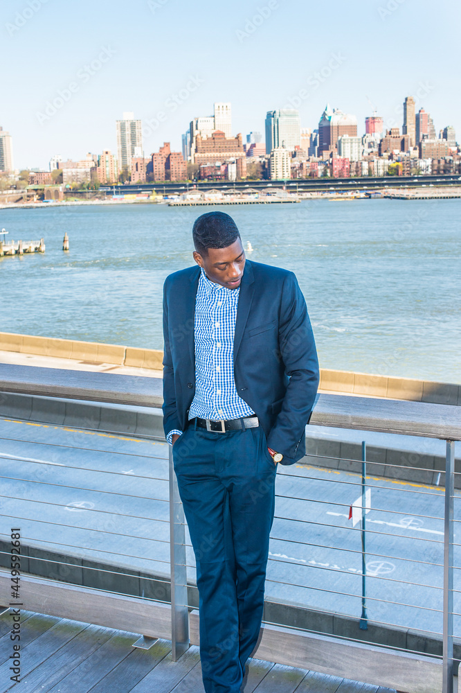 Two hands putting in pockets, lowering his head, a young black businessman is standing by the river into deeply thinking. Background is an old big city scene.. Two hands putting in pockets, lowering his head, a young black businessman is standing by the river into deeply thinking. Background is an old big city scene..