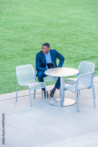 A young handsome black businessman is sitting on a chair by a green lawn, holding a small computer, looking around and waiting for you..