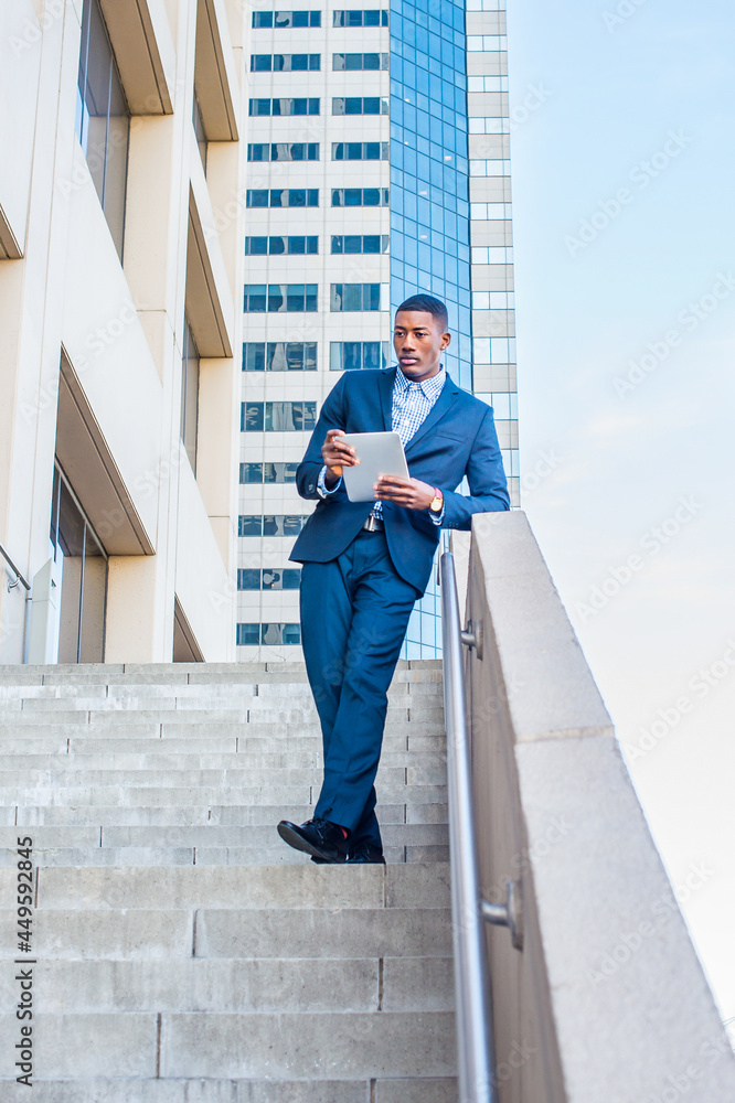 Holding a tablet computer, leaning on a railing, a young handsome black businessman is standing outside of a business building, reading, thinking and working..