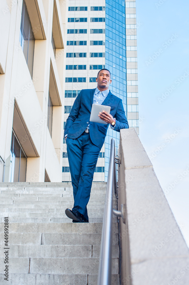 Holding a tablet computer, leaning on a railing, a young handsome black businessman is standing outside of a business building, reading, thinking and working..