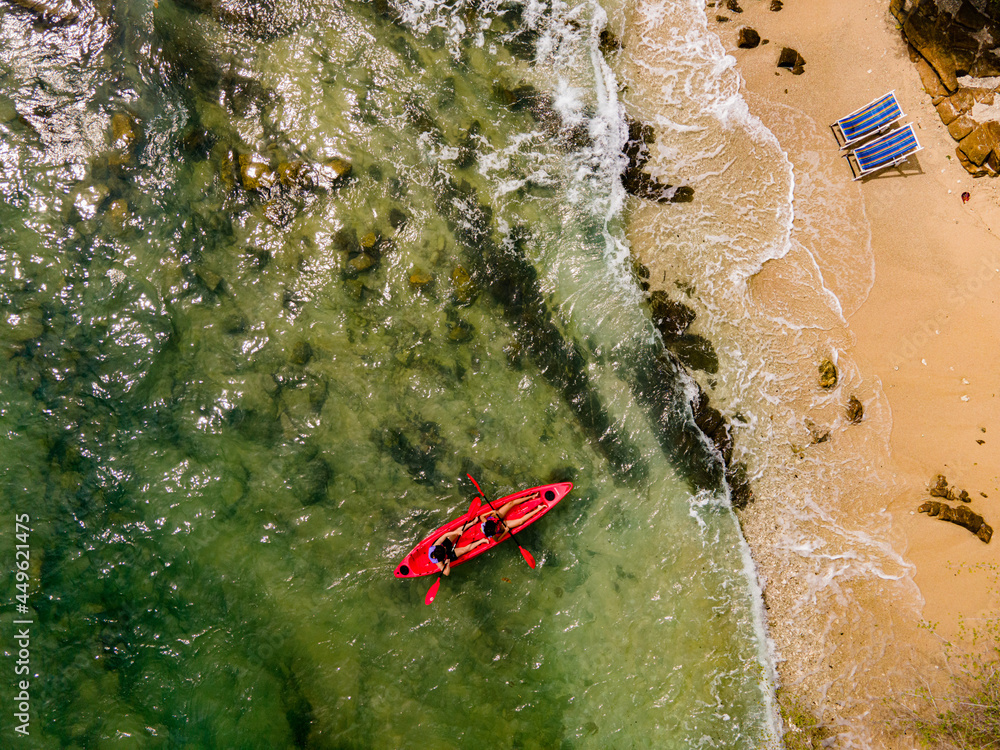 Fototapeta premium The top view of two tourist paddling their red kayak at the beatiful blue seashore and two beach chair in summer
