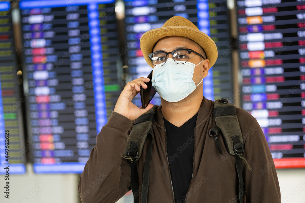 Tourist in airport terminal talking on phone to check in flight. He standing at information board. He wearing mask protect coronavirus. During the pandemic must be social distancing self quarantine.
