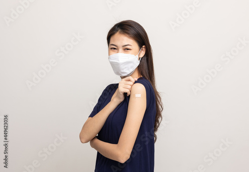 Young beautiful asian woman wearing mask and getting a vaccine protection the coronavirus. Happy female showing arm with bandage after receiving vaccination on isolated white background.