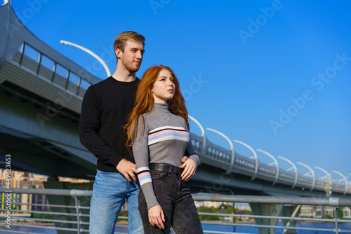 happy young couple man and woman with long red hair  stand against the background of a blue sky and a bridge in casual clothes and smile. Cheerful guy and Caucasian girl on a sunny day