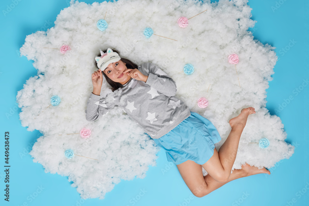 Top view of cheerful Asian woman dressed in comfortable pajama lies on fluffy cloud hears something good awakes after long night sleep poses against blue background. People and rest concept. Top view of cheerful Asian woman dressed in comfortable pajama lies on fluffy cloud hears something good awakes after long night sleep poses against blue background. People and rest concept.