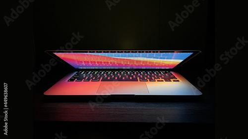 Laptop with half  lid open on a table lit with colorful desktop screen wallpaper in a dark room photo