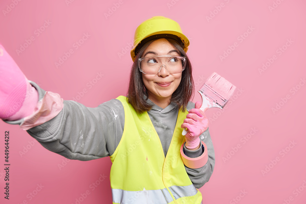 Talented master holds painting brush stretches arm refreshes walls of apartment wears protective helmet uniform and transparent glasses isolated over pink background. Renewal redecoration concept Talented master holds painting brush stretches arm refreshes walls of apartment wears protective helmet uniform and transparent glasses isolated over pink background. Renewal redecoration concept