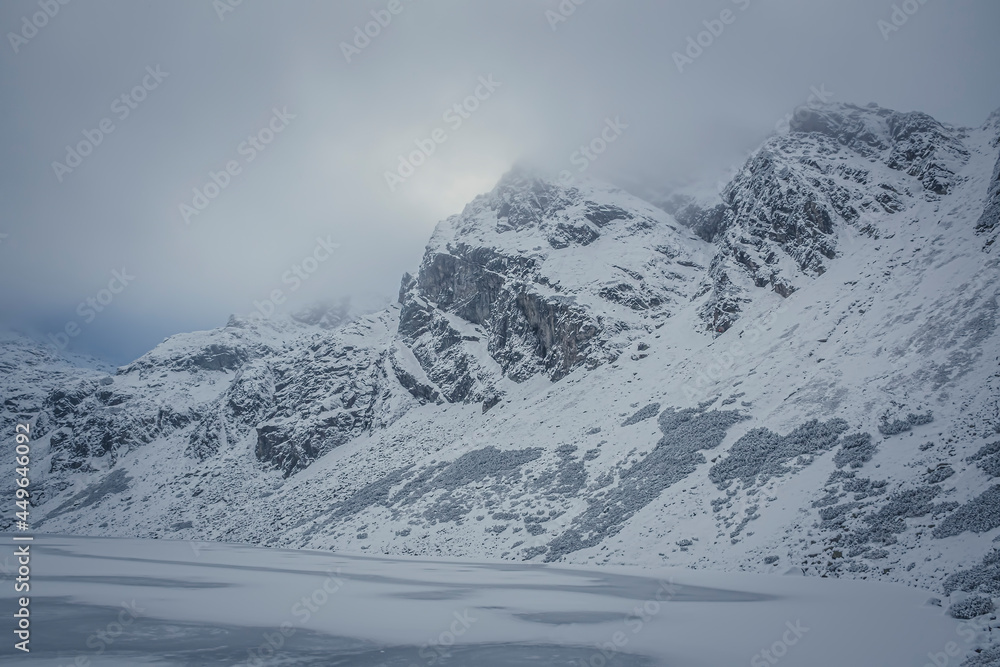 Frozen mountain lake and rocky peak, High Tatras, Poland. Czarny Staw Gąsienicowy Pond and Kościelec Summit in the clouds. Selective focus on the crags, blurred background.