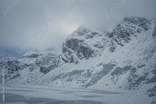 Frozen mountain lake and rocky peak, High Tatras, Poland. Czarny Staw Gąsienicowy Pond and Kościelec Summit in the clouds. Selective focus on the crags, blurred background. © juste.dcv