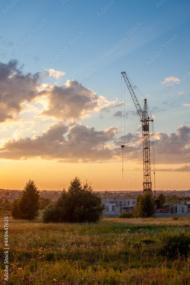 A construction site with a high-rise block under construction in an urban environment dominated by a large industrial crane, silhouetted against a beautiful cloudy sunset. construction landscape
