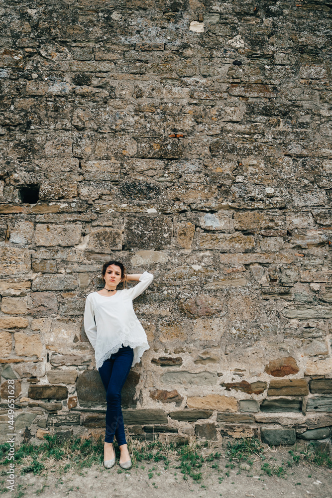 portrait of traveler girl in white dress stands at ancient stone wall
