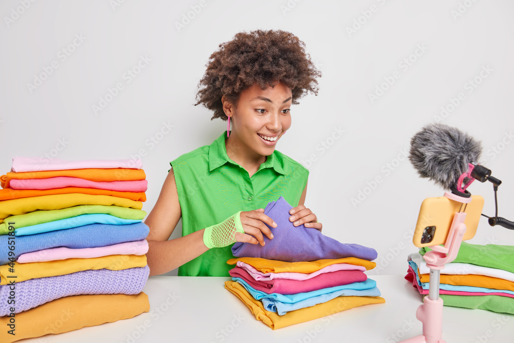 Fototapeta premium Positive woman with curly hair folds laundry in piles records live stream video being professional blogger wears green t shirt isolated over white background. People clothing and blogging concept