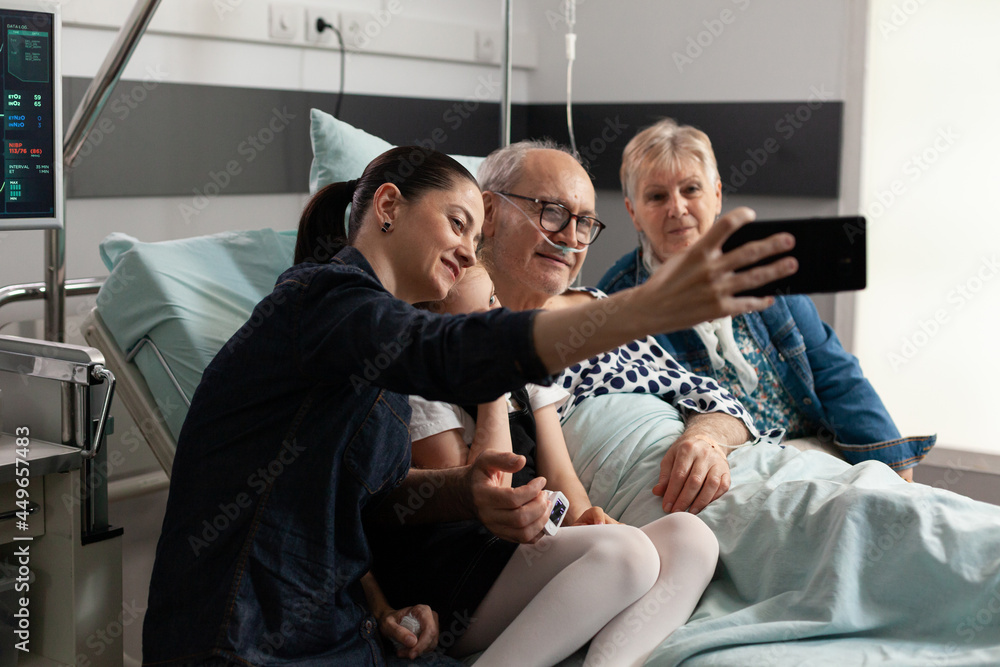 Caring family taking selfie with sick elderly grandfather using modern smartphone during clinical recovery in hospital ward. Relatives visiting senior man support him after medical surgery Caring family taking selfie with sick elderly grandfather using modern smartphone during clinical recovery in hospital ward. Relatives visiting senior man support him after medical surgery