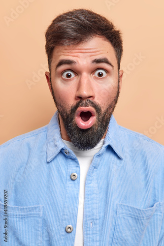 Vertical shot of bearded stupefied astonished man gasps and stares at camera with disbelief keeps mouth opened hears shocking news wears blue velvet shirt poses indoor against beige background.