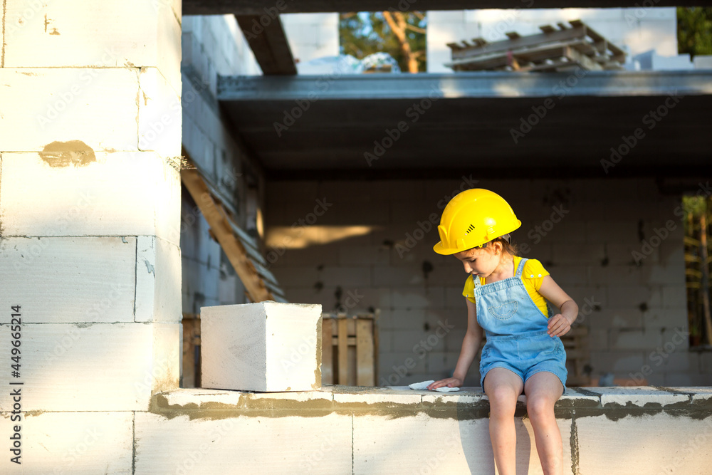 Little girl in a yellow hardhat is playing builder on the construction site of her future home. Expectation of moving, choosing a profession, children's education. Future profession, family business. Little girl in a yellow hardhat is playing builder on the construction site of her future home. Expectation of moving, choosing a profession, children's education. Future profession, family business.