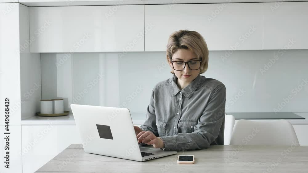 Short haired blonde businesswoman in grey shirt and glasses types on white laptop at wooden table with smartphone at home Short haired blonde businesswoman in grey shirt and glasses types on white laptop at wooden table with smartphone at home