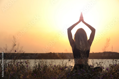 Young woman meditating near river at sunset, back view. Space for text