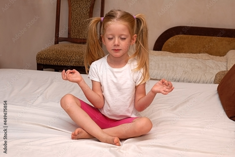 The blonde girl sits in the lotus position at home on the bed and meditates. Children's yoga. The child copies adults. The blonde girl sits in the lotus position at home on the bed and meditates. Children's yoga. The child copies adults.