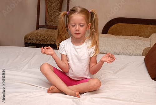 The blonde girl sits in the lotus position at home on the bed and meditates. Children's yoga. The child copies adults.